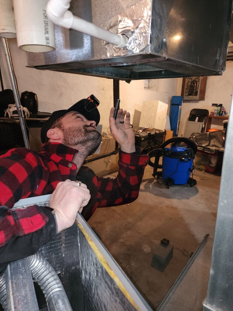 An HVAC technician inspecting an indoor furnace or air handler unit from below for Dad's Heating and Cooling in Blue Springs, MO.