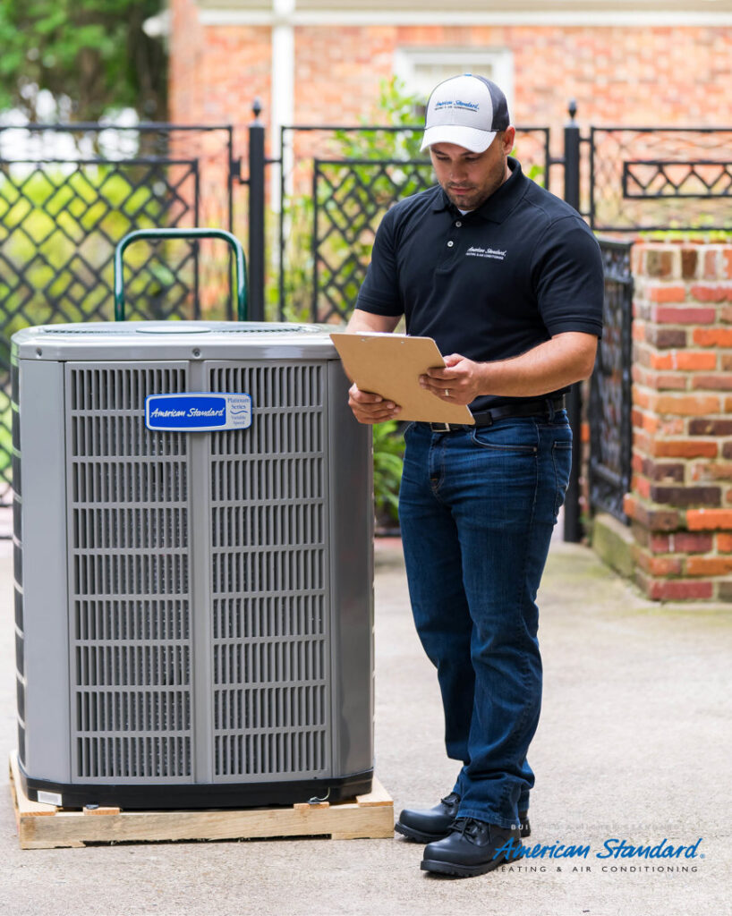 An HVAC technician in uniform inspecting an American Standard outdoor air conditioning unit for VP Supply Corporation in Rochester, NY.