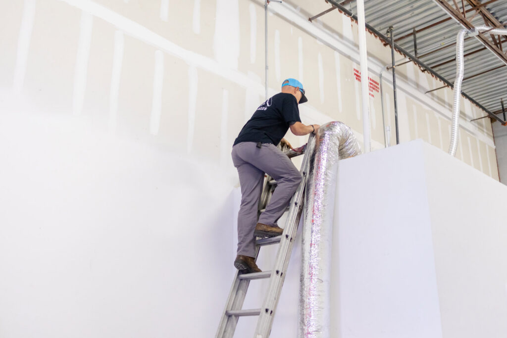 An HVAC technician on a ladder installing ductwork during a service call by Surfside Heating & Cooling in Cape Coral, FL.