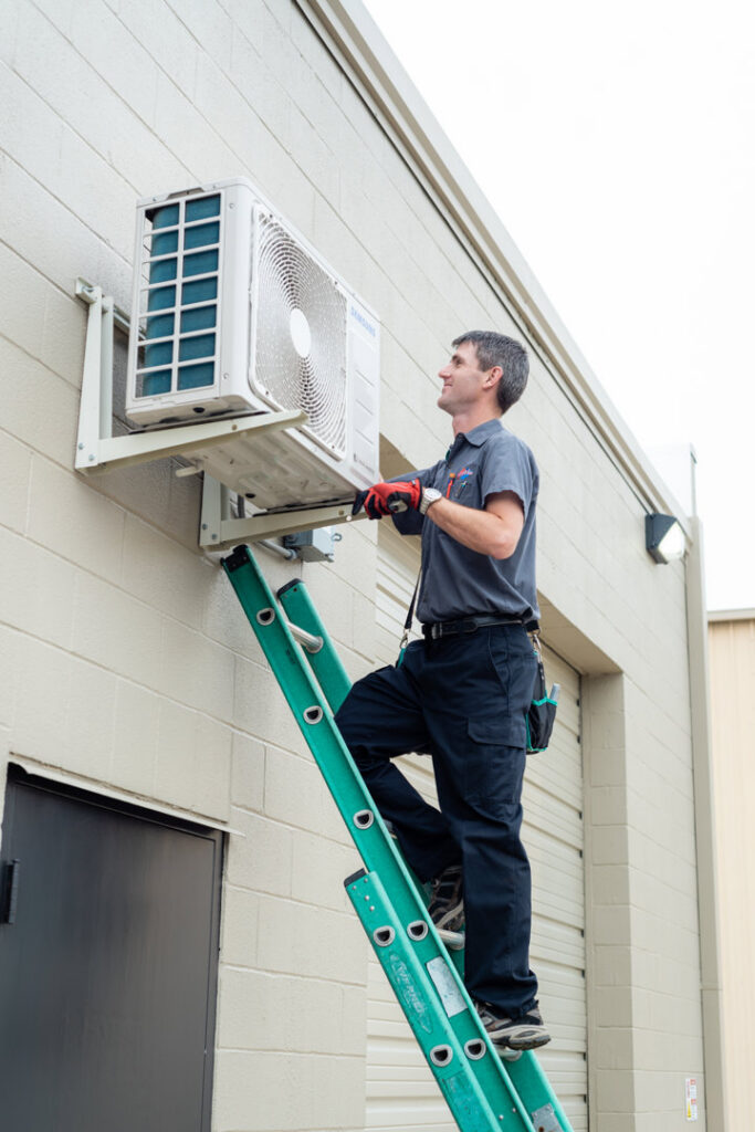 An HVAC technician on a ladder installing or servicing a mini-split outdoor unit for Calloway & Sons A/C And Heating in New Orleans, LA.