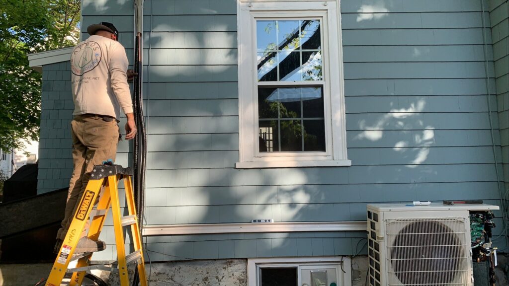 An HVAC technician installing an outdoor unit on a ladder for Atmos HVAC in Quincy, MA.