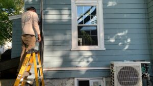 An HVAC technician installing an outdoor unit on a ladder for Atmos HVAC in Quincy, MA.