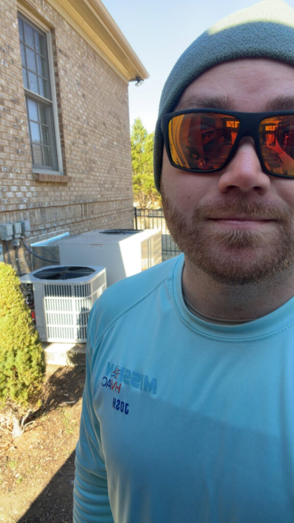 A Mission HVAC technician taking a selfie on a job site with two outdoor HVAC units in Huntsville, AL.