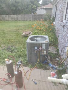 An HVAC technician from Chief/Bauer Service Experts servicing an outdoor AC unit with refrigerant tanks and gauges in Champaign, IL.