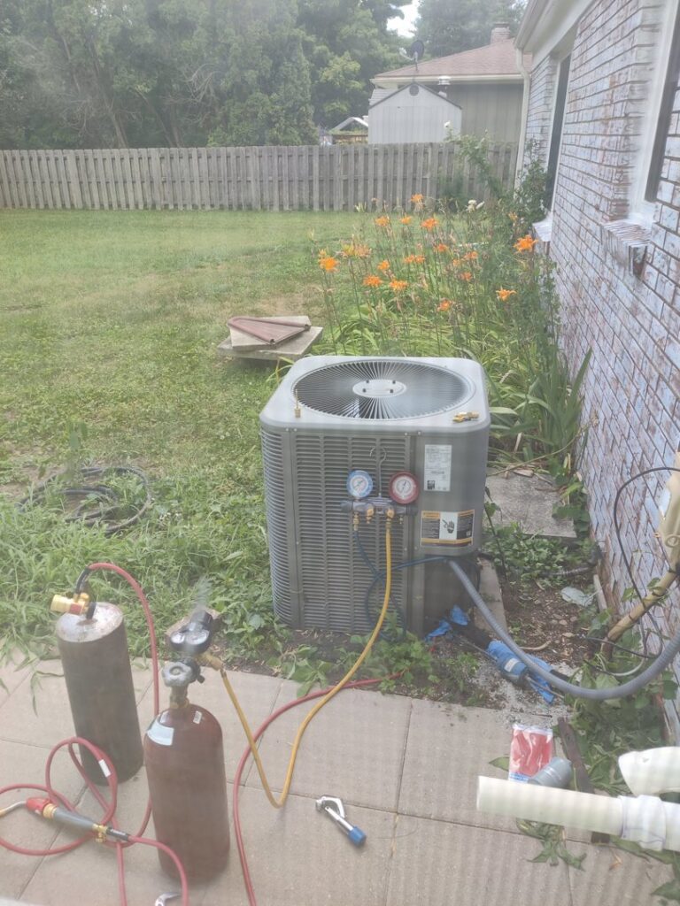 An HVAC technician from Chief/Bauer Service Experts servicing an outdoor AC unit with refrigerant tanks and gauges in Champaign, IL.