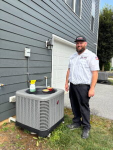 An HVAC technician from One Hour Heating & Air Conditioning of Lancaster, PA, standing next to an outdoor AC unit with cleaning supplies.