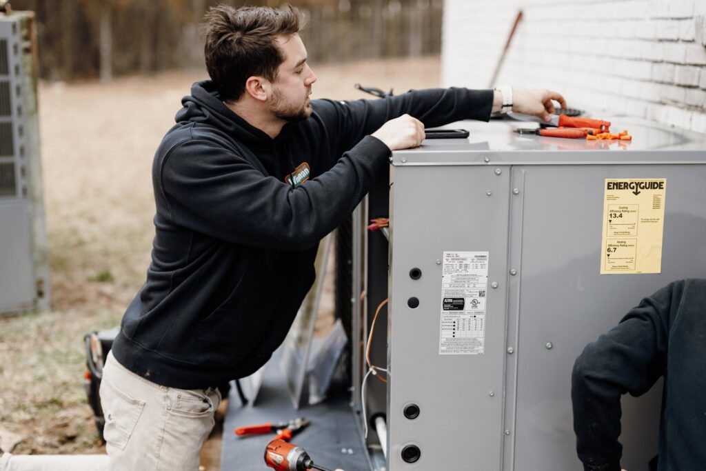 An HVAC technician performing service on an outdoor air conditioning unit with tools nearby for Wingman Heating + Cooling in Auburn, AL.