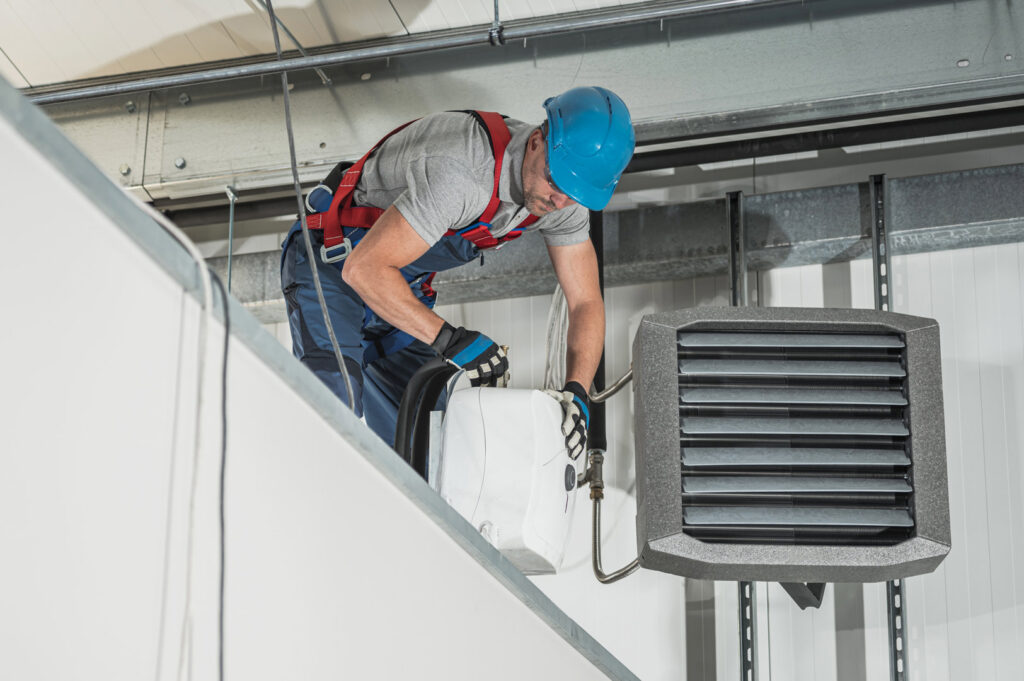 An HVAC technician in a hard hat servicing an overhead commercial heating unit, performed by Jons-Air in Carmel, IN.