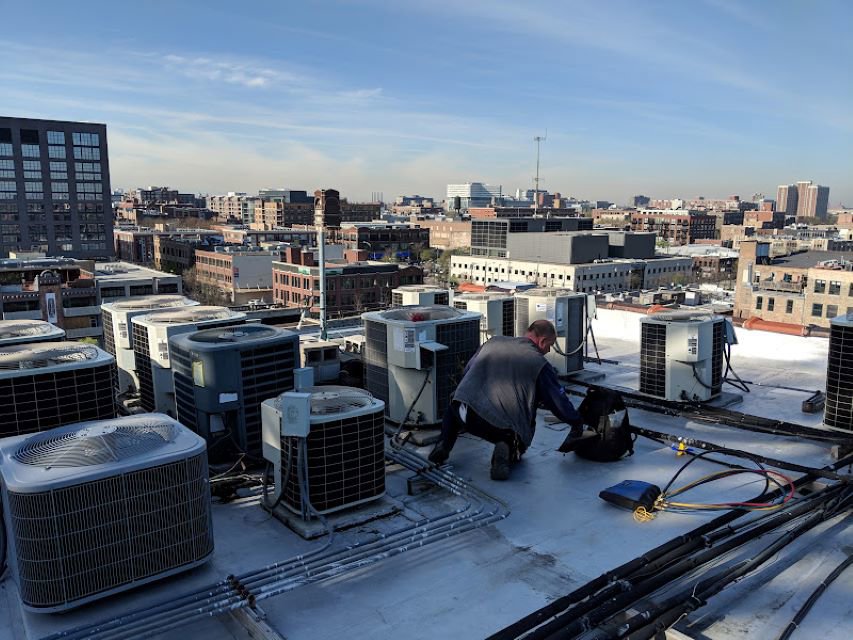 An HVAC technician from Deljo Heating, Cooling & Plumbing servicing multiple outdoor AC units on a commercial rooftop in Chicago, IL.