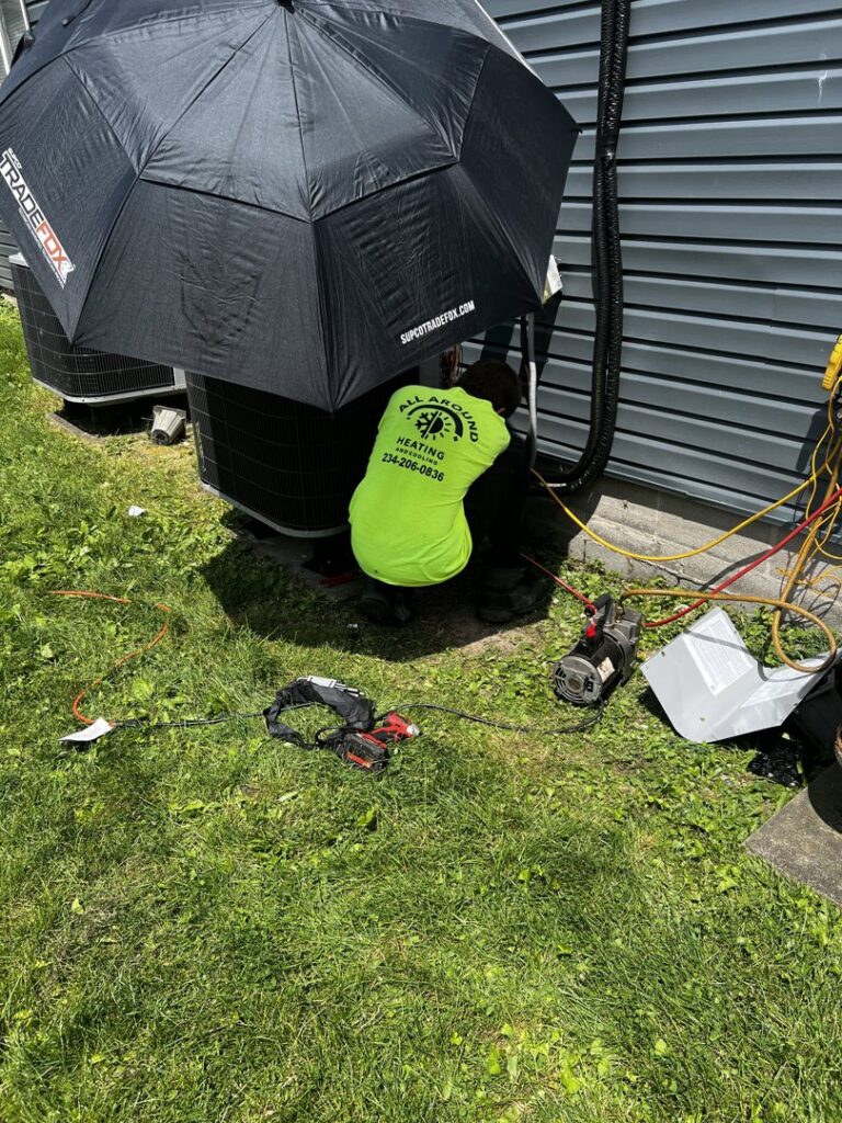 An HVAC technician from All Around Heating & Cooling servicing an outdoor air conditioning unit under an umbrella in Akron, OH.
