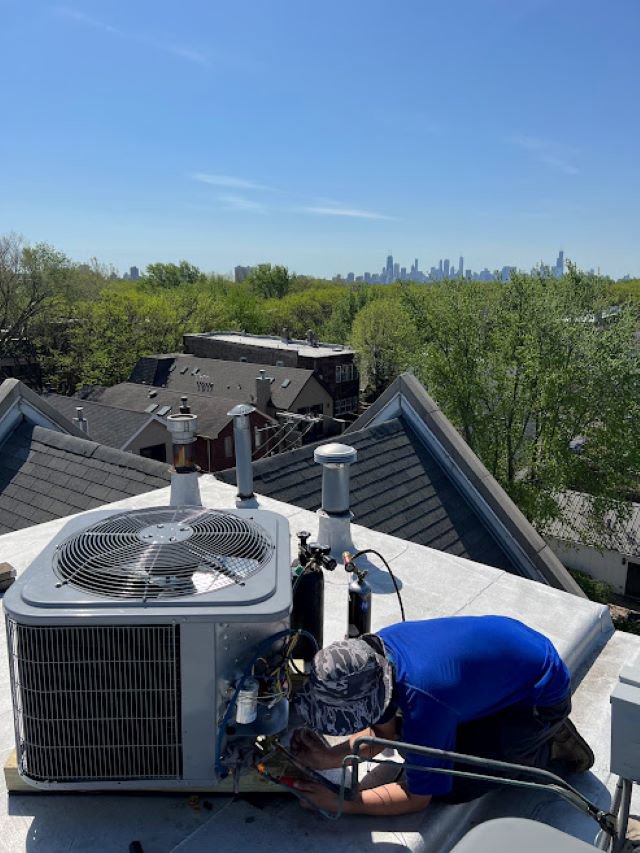 An HVAC technician from Deljo Heating, Cooling & Plumbing servicing a rooftop AC unit with the Chicago skyline in the background.