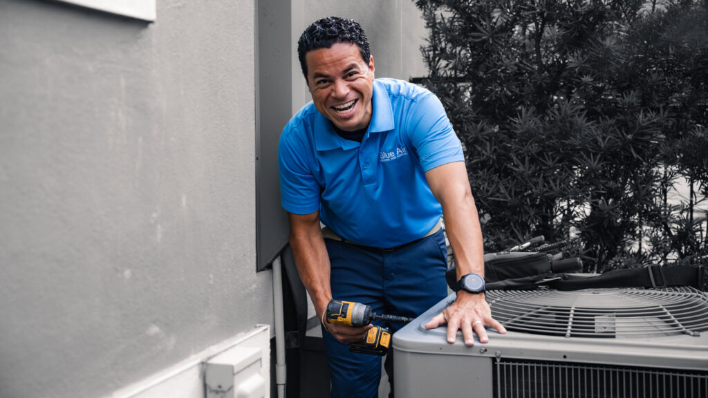 A smiling HVAC technician holding a drill next to an outdoor AC unit for Blue Air Heating and Cooling in Orlando, FL.