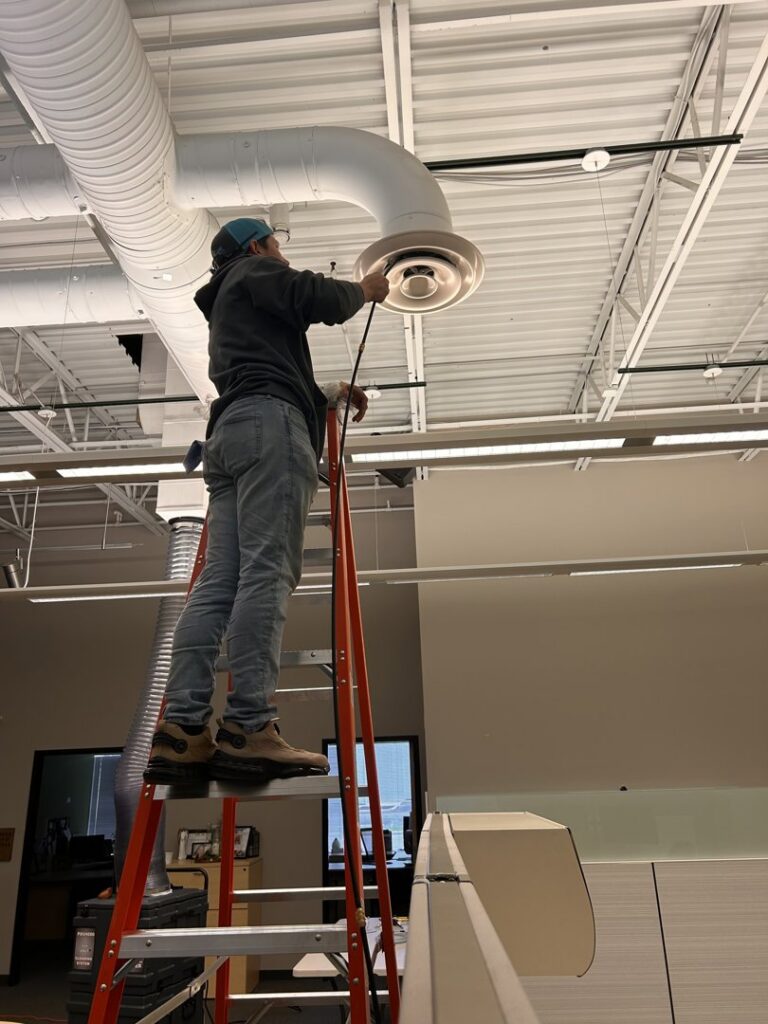 An HVAC technician on a ladder performing service on a ceiling duct and vent for Varsity Zone HVAC of Fishers, IN.