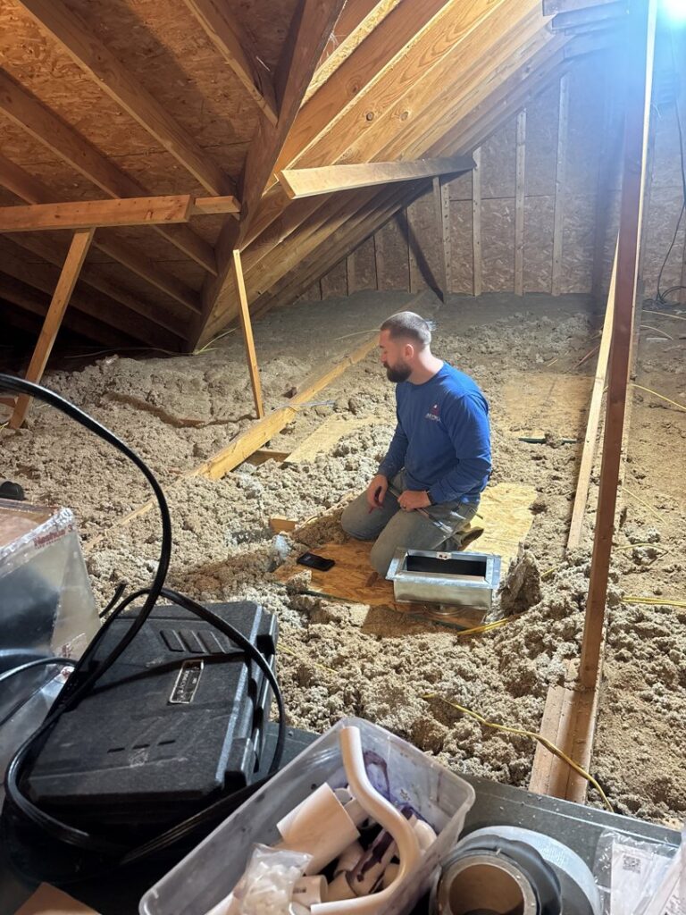 An HVAC technician kneeling amidst attic insulation, working on HVAC components for Dewees HVAC in Conway, AR.