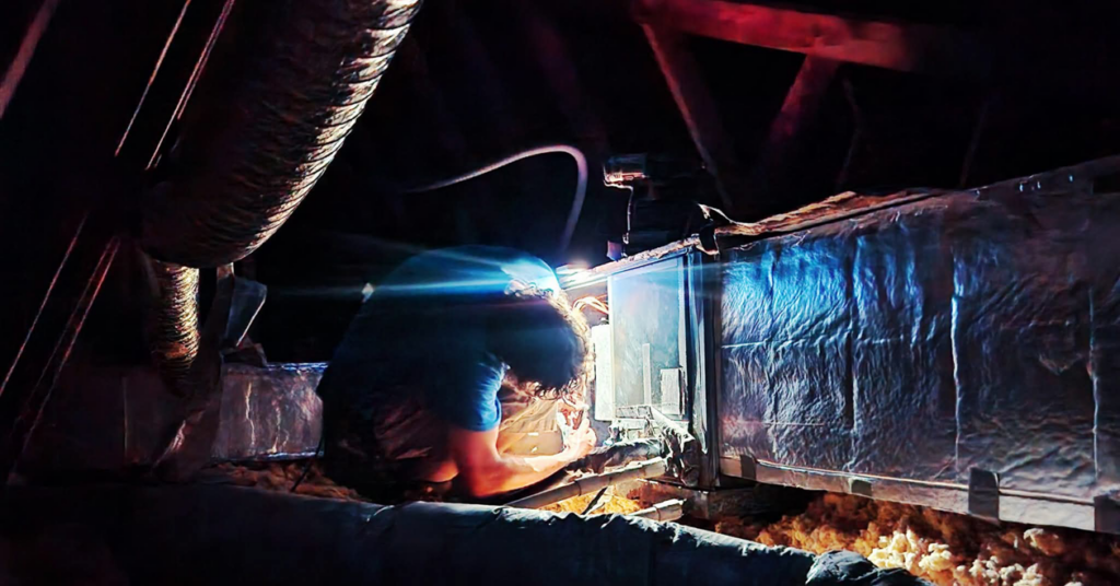 An HVAC technician working on an air handler unit in an attic, surrounded by ductwork, for Whittington HVAC in King, NC.