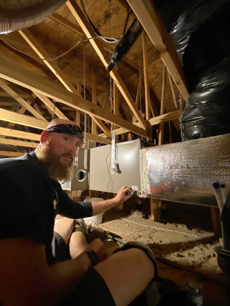 An HVAC technician from TK Heating and Air, PLLC working on ductwork and an HVAC unit in an attic in Queen Creek, AZ