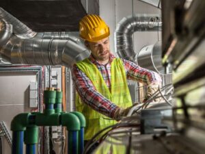 An HVAC technician in a hard hat and safety vest working on ductwork and wiring for Garden City Plumbing & Heating in Missoula, MT