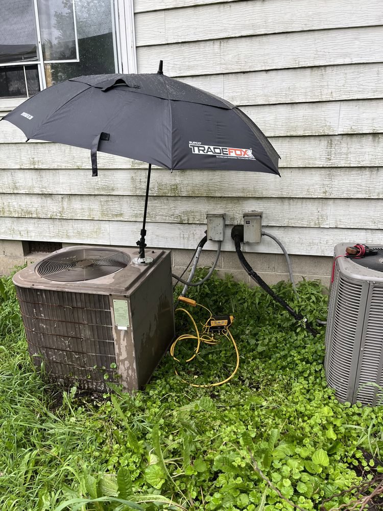 An HVAC technician's setup with an umbrella and tools next to two outdoor air conditioning units, showing a service call by Greenley Heating & Cooling in Waterloo, IA