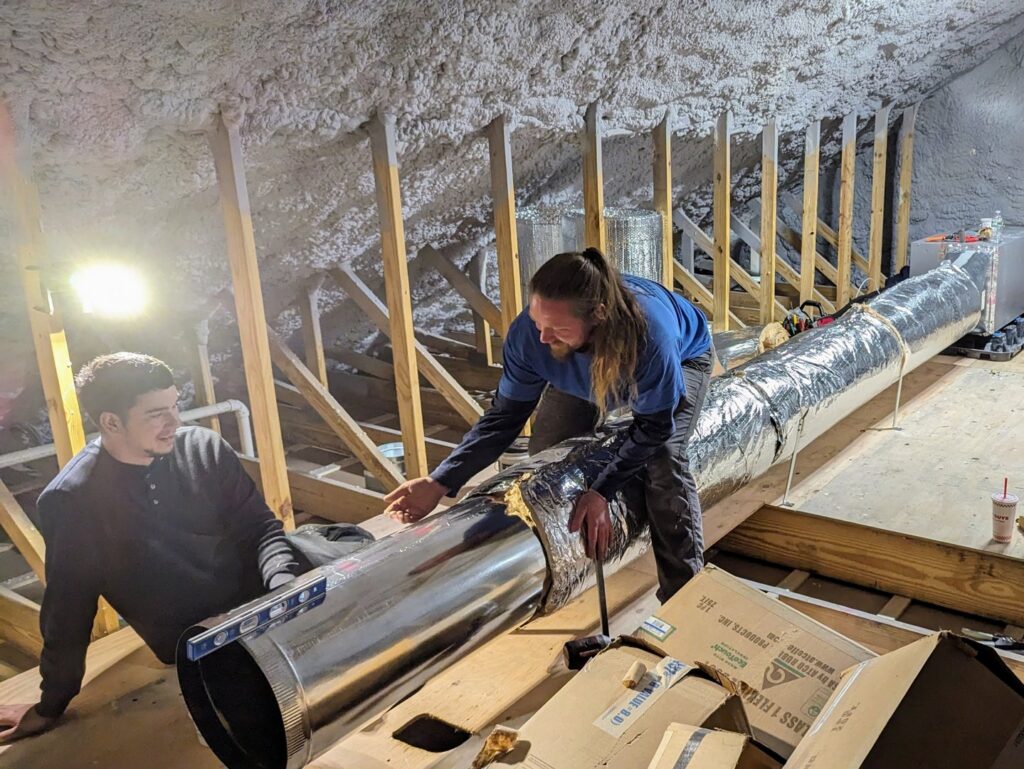 Two HVAC technicians installing large ductwork in an attic space for WARM Global HVAC in East Providence, RI