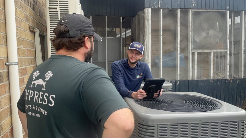 HVAC technicians discussing work near outdoor condenser units, providing service from Grand Slam Heating & Air in Baton Rouge, LA.