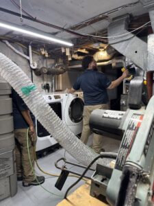 HVAC technicians performing duct cleaning and maintenance in a basement utility room for Aspen Air Duct Cleaning in Methuen, MA.