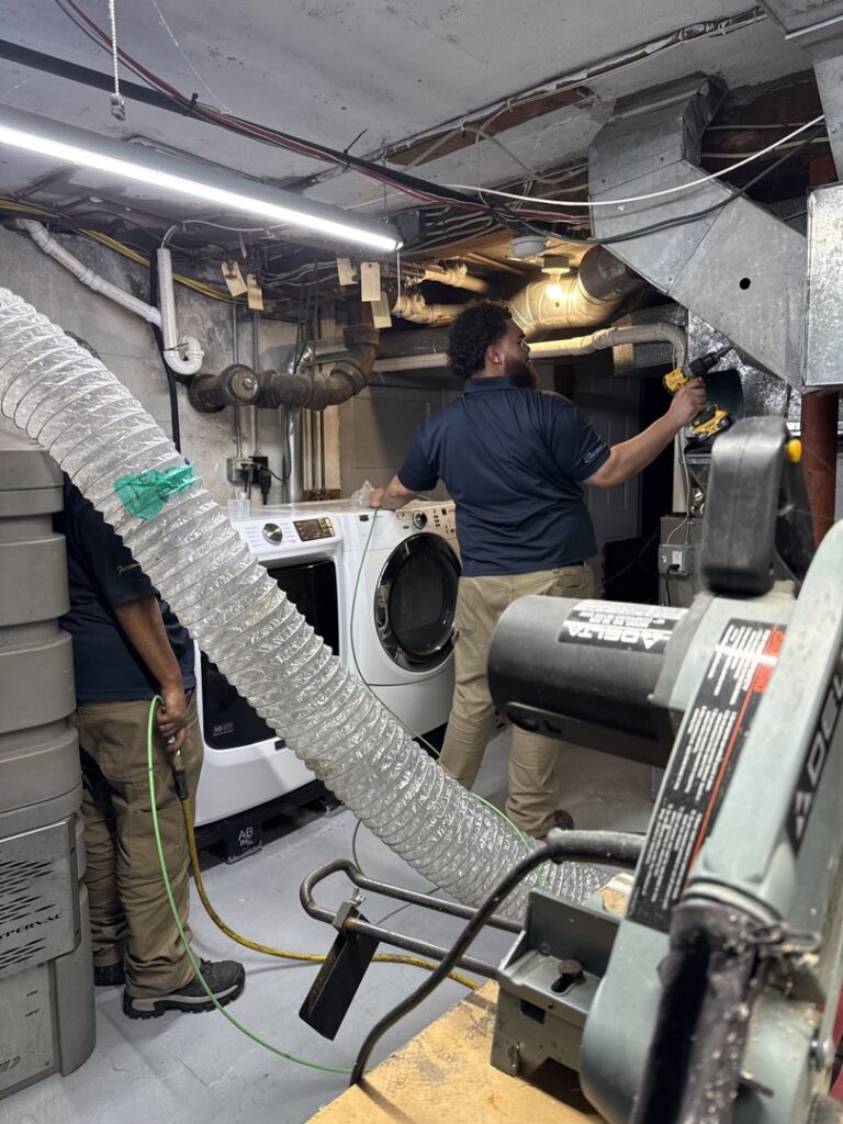 HVAC technicians performing duct cleaning and maintenance in a basement utility room for Aspen Air Duct Cleaning in Methuen, MA.