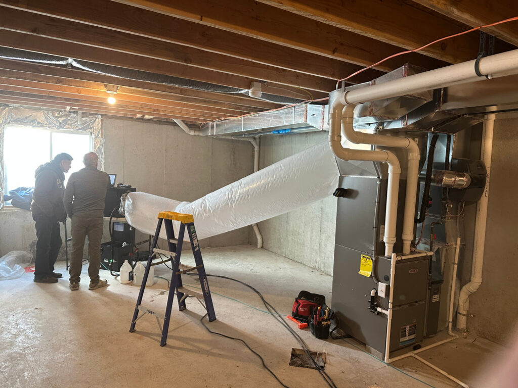 Two HVAC technicians working on a furnace and duct system in a basement for Green Seal Energy in Overland Park, KS.