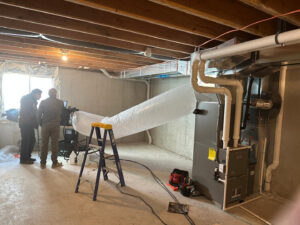 Two HVAC technicians working on a furnace and duct system in a basement for Green Seal Energy in Overland Park, KS.