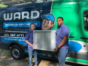 Two HVAC technicians holding an indoor AC unit in front of their service van for Ward Mechanical Contractors in Denham Springs, LA.