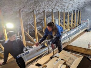 Two HVAC technicians from WARM Global HVAC installing new ductwork in an attic space in East Providence, RI.