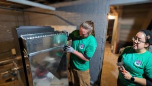 Two HVAC technicians from WARM Global HVAC installing new ductwork in a basement or utility room in East Providence, RI.