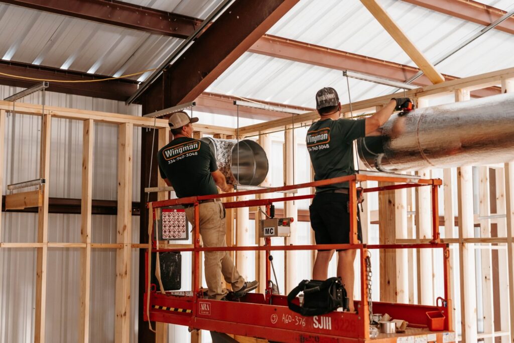Two HVAC technicians on a scissor lift installing large ductwork in a commercial building for Wingman Heating + Cooling in Auburn, AL.