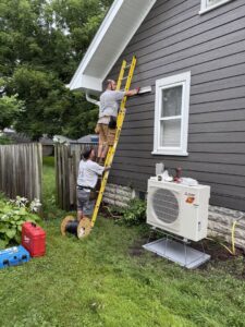 Two A&E Heating & Cooling technicians installing a Mitsubishi mini-split unit on a house exterior in Fort Worth, TX.