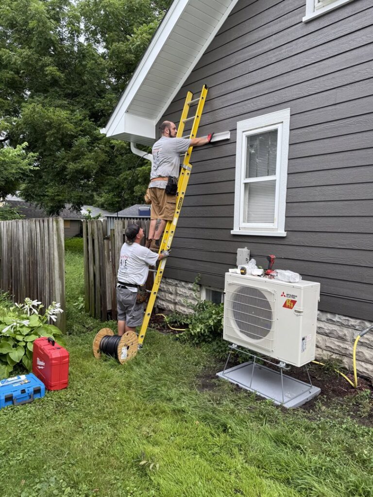 Two A&E Heating & Cooling technicians installing a Mitsubishi mini-split unit on a house exterior in Fort Worth, TX.