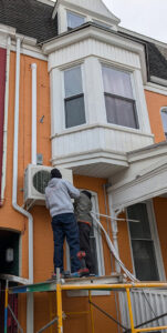 HVAC technicians installing an outdoor mini-split condenser unit on a building for Strine's Heating & A/C in York, PA.