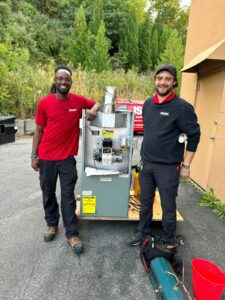 Two HVAC technicians standing with a new furnace ready for installation by Isaac Heating & Air Conditioning in Rochester, NY.