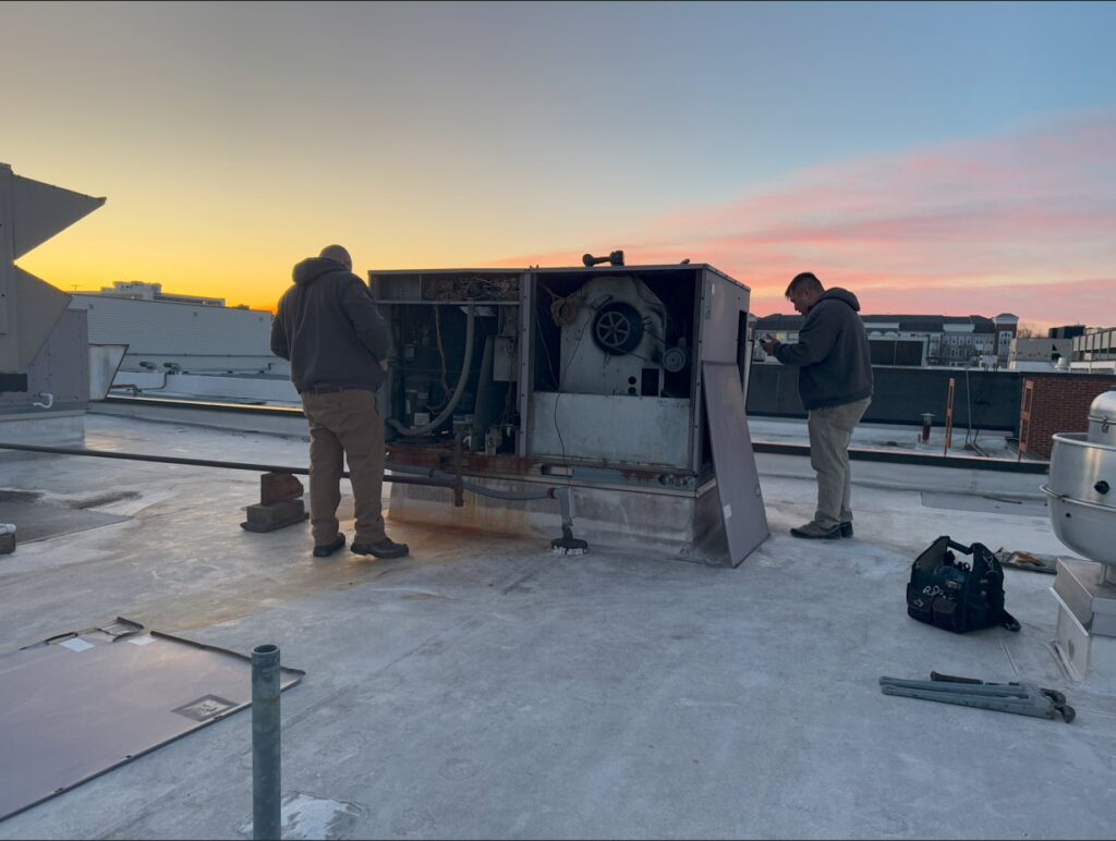 Two HVAC technicians repairing an older rooftop unit at sunset for APEX HVAC Solutions LLC in Frederick, MD.