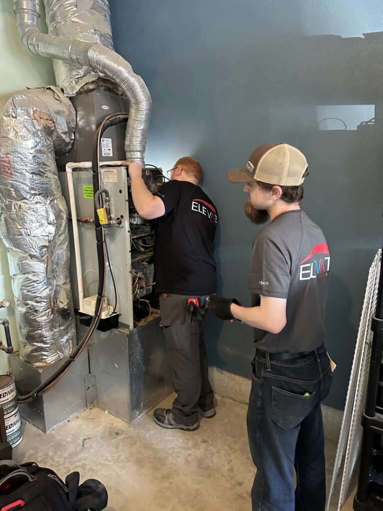 Two HVAC technicians servicing an indoor furnace unit at Elevate Heating & Cooling in Hillsboro, OR.