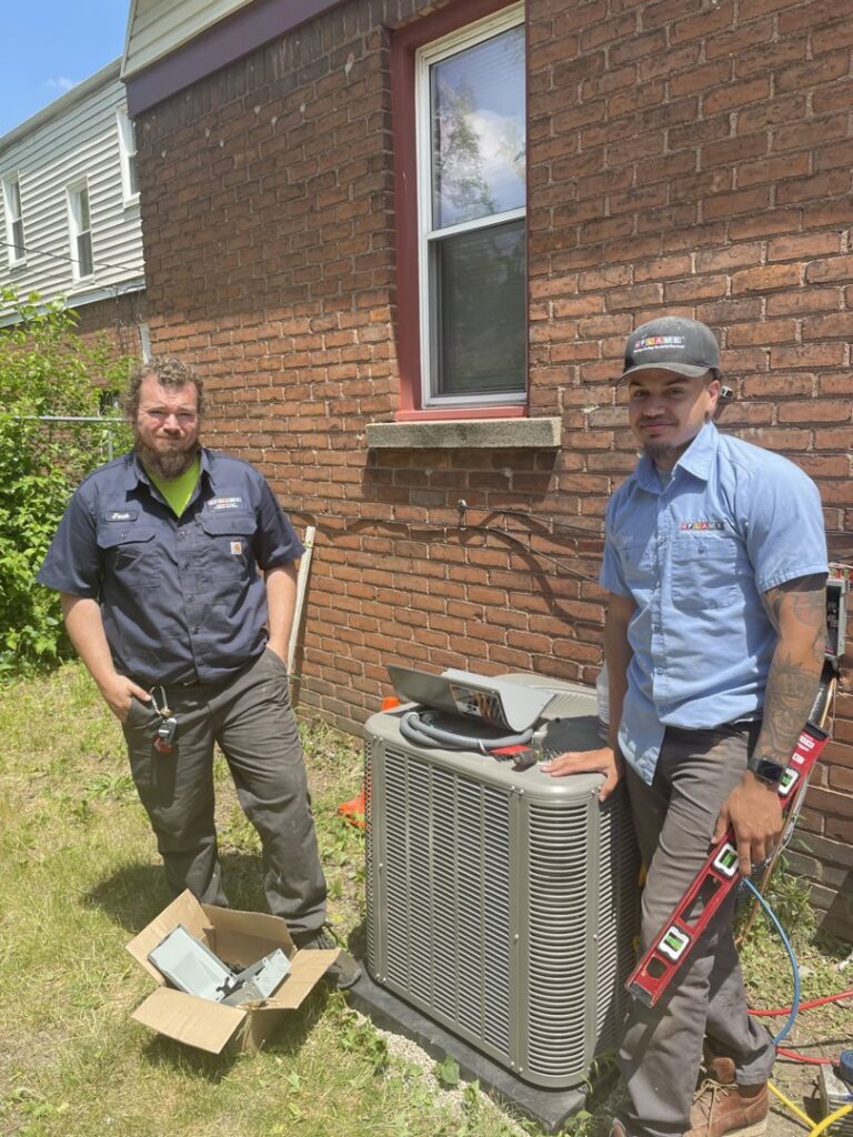 Two FLAME Heating, Cooling, Plumbing & Electrical technicians working on an outdoor AC unit in Warren, MI.