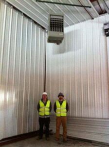Two HVAC technicians in safety vests standing with a large overhead heating unit in a commercial building, installed by KATS Heating & Cooling in La Grange Park, IL.