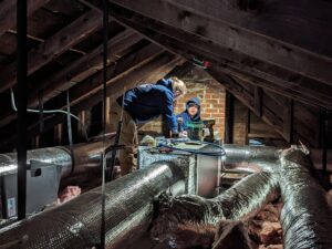 Two HVAC technicians from WARM Global HVAC working on ductwork and an air handler in an attic in East Providence, RI.