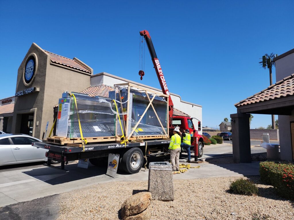 A crane lifting a large, crated HVAC unit from a delivery truck, part of a commercial installation by State 48 Home Comfort LLC in Peoria, AZ.