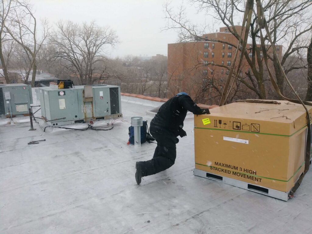 A worker on a snowy rooftop next to a large packaged HVAC unit for installation by Bean'z Heating and Cooling in Chicago, IL.