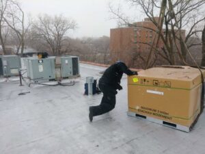 A worker on a snowy rooftop next to a large packaged HVAC unit for installation by Bean'z Heating and Cooling in Chicago, IL.