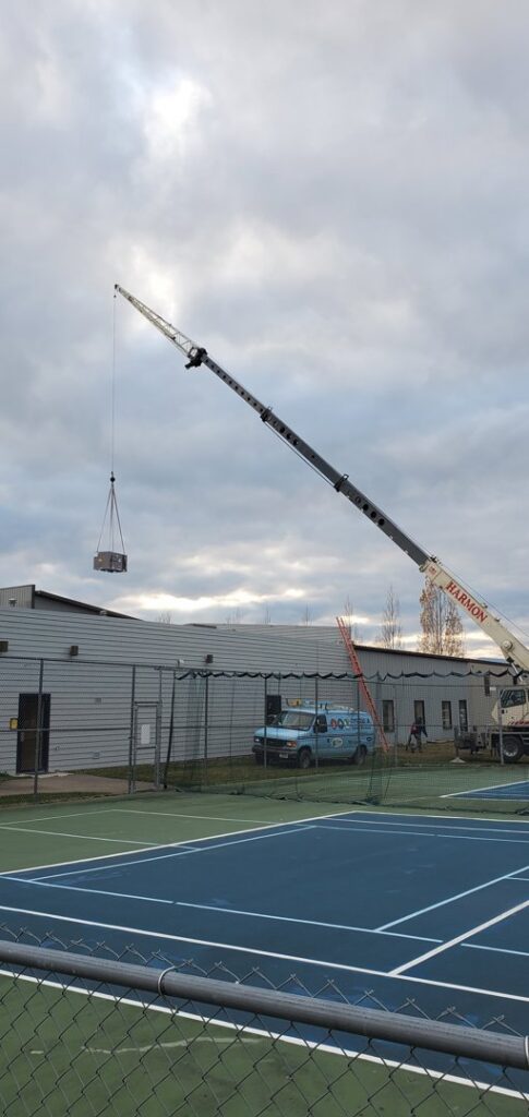 A large HVAC unit is carefully lifted by a crane over a tennis court towards a building by Comfort Systems USA Temp Right Service in Kalispell, MT.