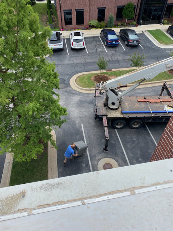 A worker transporting an HVAC unit with a crane truck in the background, handled by Dowd Heat & Air in Tulsa, OK.