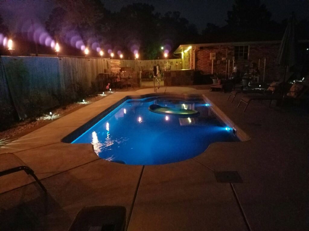 An illuminated freeform swimming pool at night with blue lights and tiki torches by Central Pools, Inc. in Baton Rouge, LA