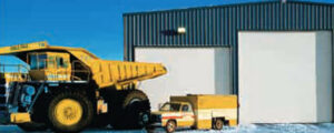 An industrial building with a large white garage door next to a yellow dump truck by Overhead Door Company of Knoxville in Knoxville, TN