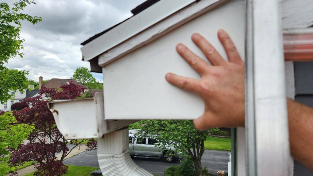 A hand inspecting damaged soffit on a house, identifying potential wildlife entry points for Armstrong's Wildlife Solutions in Rochester, NY.