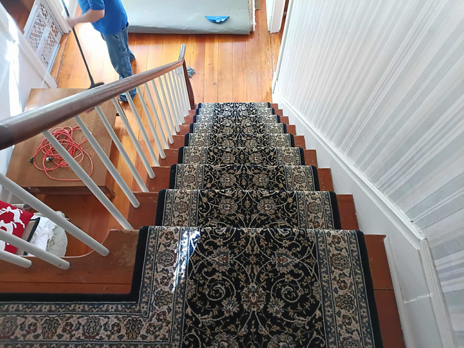 A professional installing a dark patterned carpet runner on a staircase for Onondaga Flooring in Syracuse, NY.